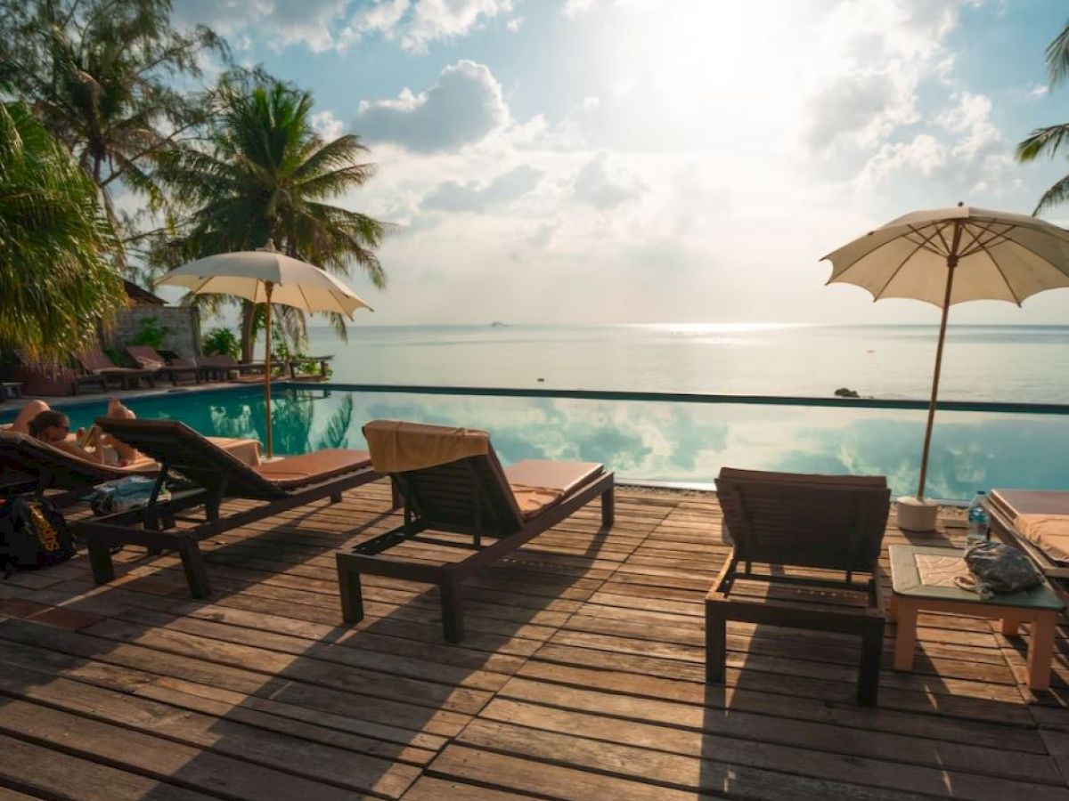 A tropical poolside scene with lounge chairs, umbrellas, and palm trees overlooking a serene body of water under a bright sky.