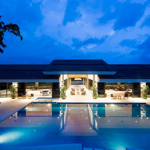 The image shows a modern building with a large, illuminated pool in the foreground, set against a twilight sky.