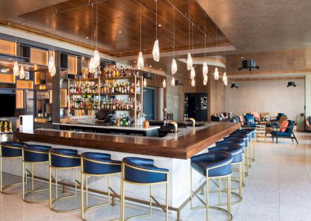 The image shows a modern bar with blue barstools, pendant lighting, and a well-stocked back bar in a spacious, stylish setting.