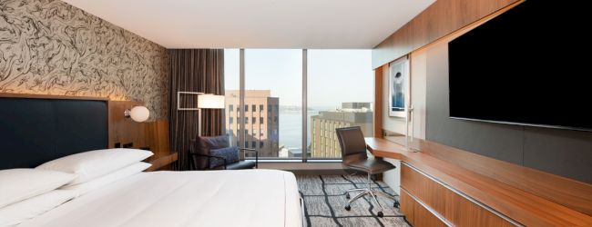 A modern hotel room with a large bed, wall-mounted TV, desk, chair, and a window with a city view, featuring contemporary decor.
