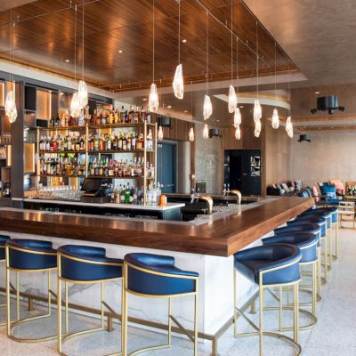 This image shows a modern bar area with a central counter, blue stools, stylish lighting, and shelves with bottles, creating a cozy atmosphere.