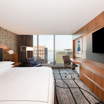 A modern hotel room with a large bed, wall-mounted TV, desk, and floor-to-ceiling windows offering a city view.