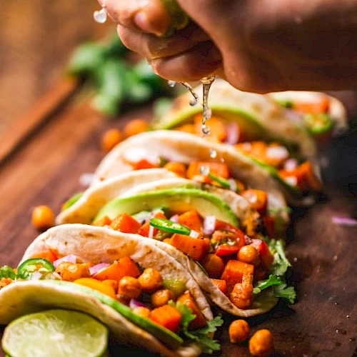 Tacos with veggies, chickpeas, avocado, onion; lime being squeezed over them on a wooden board.