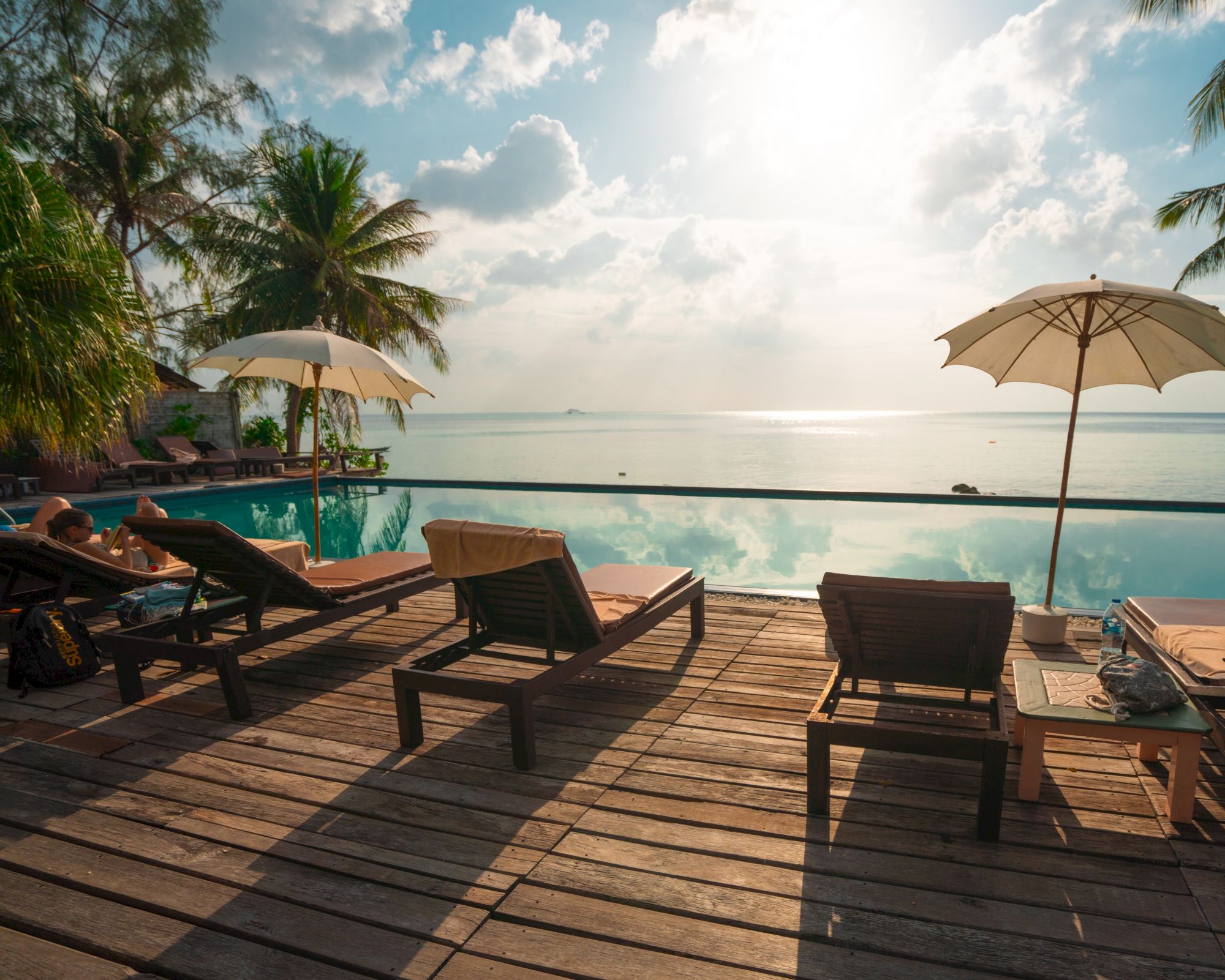 Lounge chairs and umbrellas by a poolside with a view of the ocean and a beautiful sky in the background.