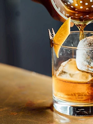 A classic cocktail being poured into a glass with ice and an orange twist garnish on a bar counter.