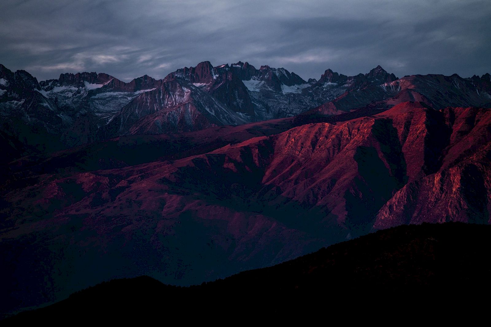 A mountain range at dusk with reddish hues illuminating the rugged peaks under a cloudy sky.