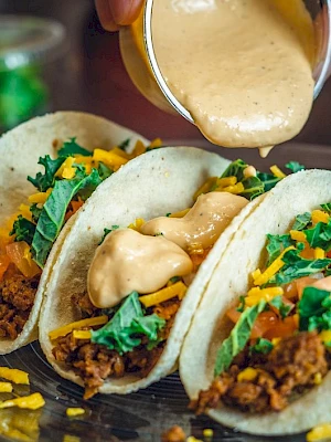 Three tacos topped with cheese sauce, filled with ground meat, lettuce, cheese, and tomatoes are on a plate.