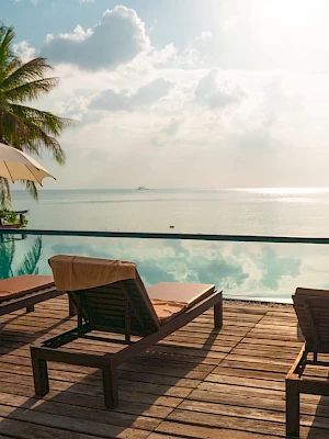 A tropical beach scene with lounge chairs, umbrellas, and palm trees by an infinity pool overlooking a tranquil ocean at sunset.