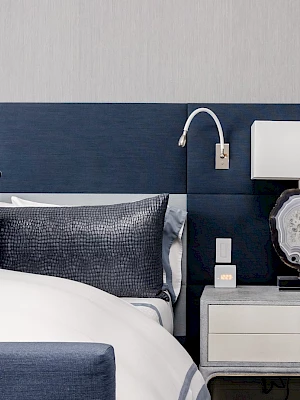 A modern bedroom features a bed with blue pillows, a navy headboard, a white two-drawer nightstand, a lamp, and a wall-mounted reading light.