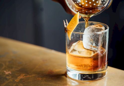 A cocktail is being poured into a glass with ice and a citrus peel garnish on a bar counter.