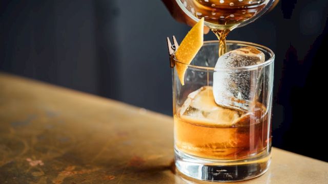 A cocktail is being poured into a glass with ice and a citrus peel garnish on a bar counter.