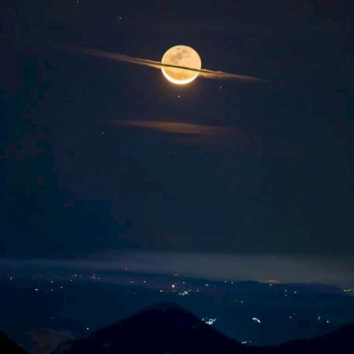The image shows the moon with clouds creating the illusion of Saturn's rings above a dark landscape with distant lights.