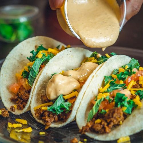 Three tacos with meat, cheese, and greens being topped with sauce on a plate, creating a delicious presentation.