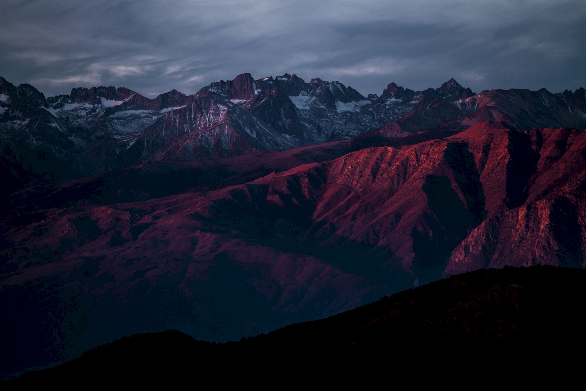A mountain range with a dramatic red hue under a cloudy sky at dusk or dawn.