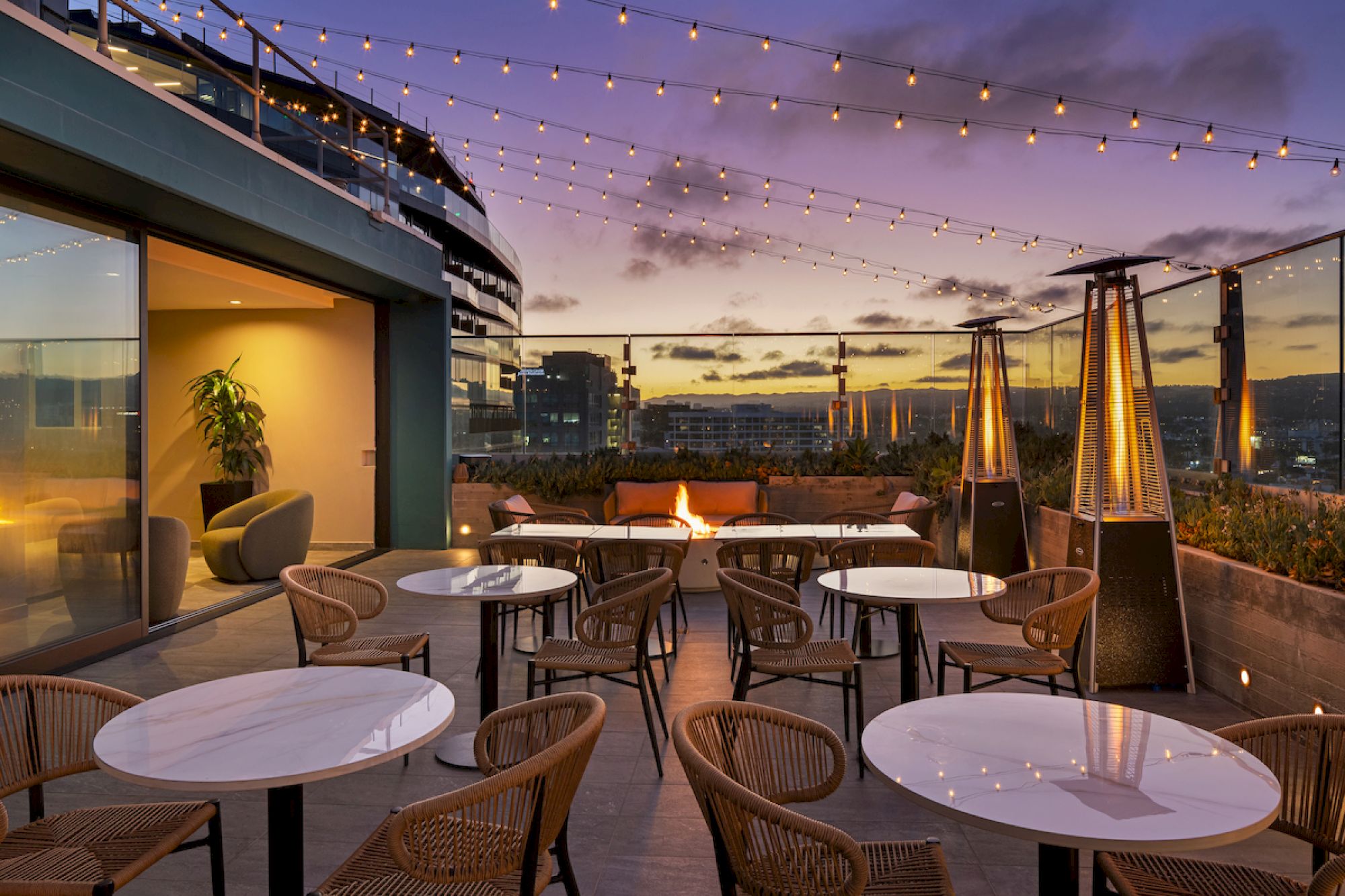 A rooftop terrace with round tables and wicker chairs, surrounded by heaters and string lights, overlooks a sunset cityscape.