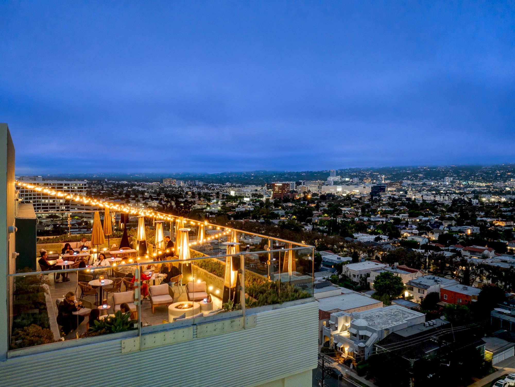 A rooftop bar is lit with string lights, overlooking a sprawling cityscape at dusk, creating a cozy and vibrant ambiance.