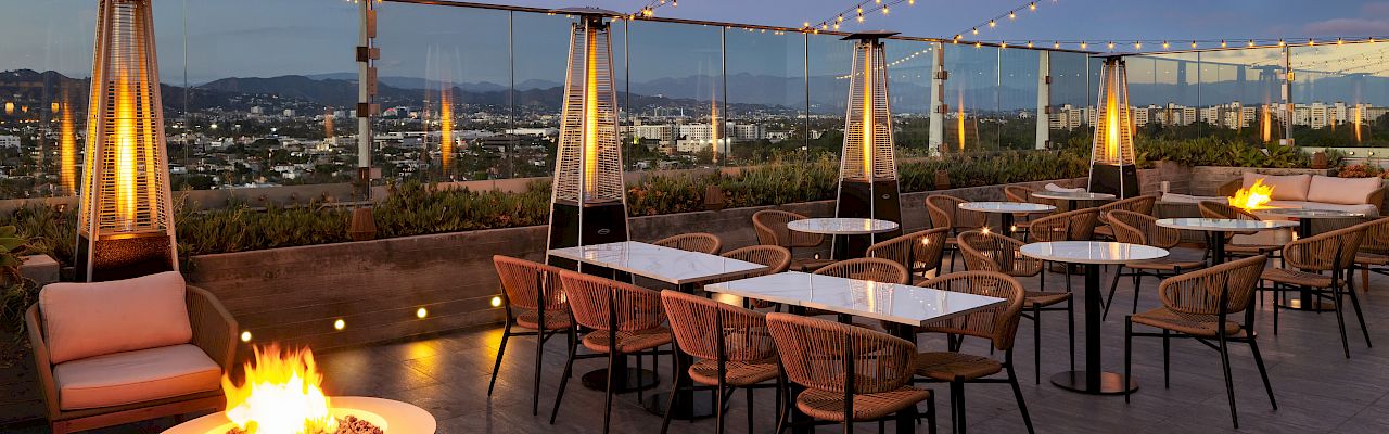 A stylish rooftop patio with tables, chairs, string lights, tall heaters, and a fire pit, overlooking a city skyline at dusk.
