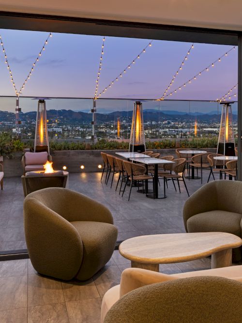 The image shows a stylish rooftop patio with comfortable seating, tables, and string lights under a twilight sky with a scenic view.