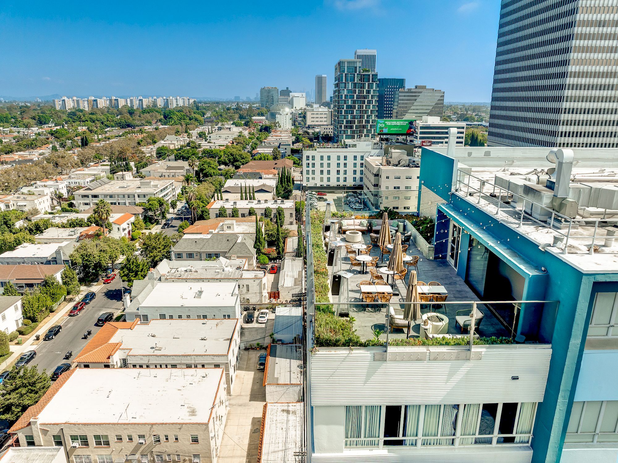 A rooftop with seating and greenery overlooks a cityscape with tall buildings and a clear blue sky in the background.