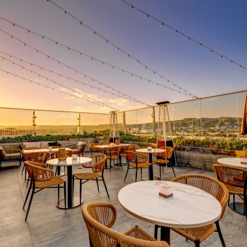 A rooftop patio with wicker chairs and round tables under string lights, overlooking a scenic sunset view.