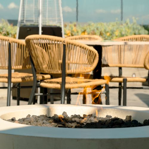A patio setting with wicker chairs and a circular fire pit surrounded by stones, against a backdrop of greenery and a tall outdoor heater.