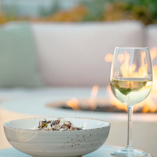 A bowl of food next to a glass of white wine sits on a small table, with a blurred background featuring a fire pit.