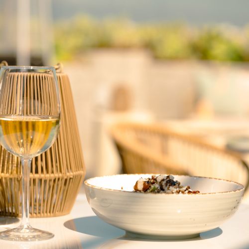 A glass of white wine and a bowl of food on a sunlit table, with a woven lamp beside them outdoors.