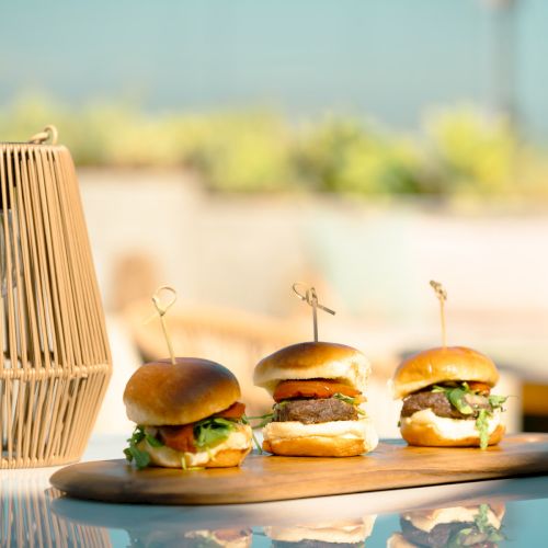 Three gourmet sliders with beef patties and fresh toppings are neatly arranged on a wooden board outdoors, accompanied by a decorative lantern.