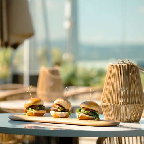 Three sliders on a plate sit on a round table with a wicker lamp, surrounded by outdoor seating in a sunny setting.