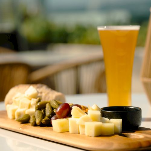 A wooden platter with cheese cubes, pickles, grapes, and bread sits alongside a glass of beer on a sunny outdoor table.
