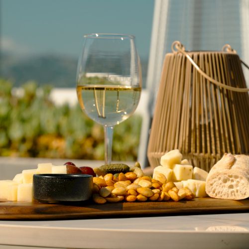 A cheese platter with various cheeses and bread next to a glass of white wine and a decorative lantern on an outdoor table.