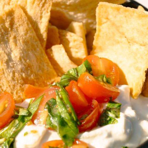 A bowl of pita chips with a creamy dip topped with cherry tomatoes and chopped green herbs.