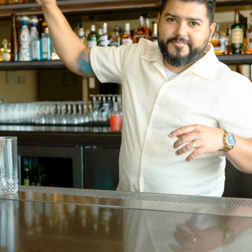 A bartender with a beard is shaking a cocktail shaker in a bar, with bottles and glasses in the background.