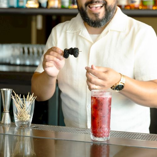 A person is garnishing a red cocktail with blackberries at a bar, smiling, with bottles and tools in the background.