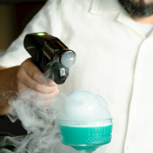 A person uses a device to create smoke bubbles over a turquoise drink in a fancy glass, adding an artistic touch to the presentation.