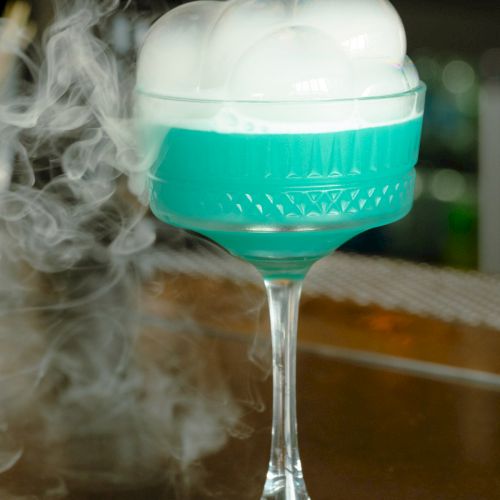A cocktail in a tall glass with blue liquid, topped with foam and surrounded by smoke, likely from dry ice.