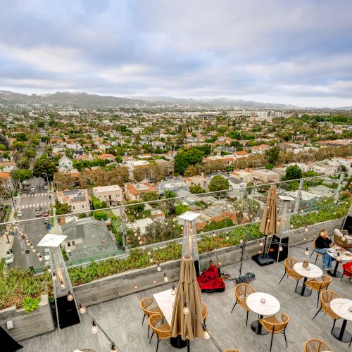 A rooftop terrace with tables, chairs, and umbrellas overlooks a sprawling urban landscape under a cloudy sky.