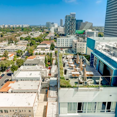Image shows a cityscape with a rooftop terrace featuring seating areas and greenery, surrounded by buildings under a clear blue sky.