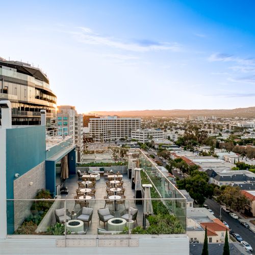A rooftop terrace with seating and tables, overlooking a cityscape under a blue sky, with buildings and trees in the distance.