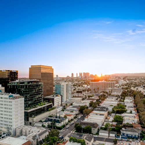A city skyline at sunset with tall buildings, a vibrant blue sky, and the sun setting on the horizon.