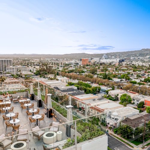 A rooftop terrace with tables and chairs overlooks a sprawling urban landscape with distant mountains under a clear blue sky.