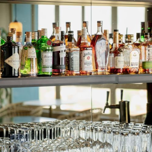 A bar shelf with various liquor bottles, including gin and whiskey, and a row of neatly arranged glasses below.