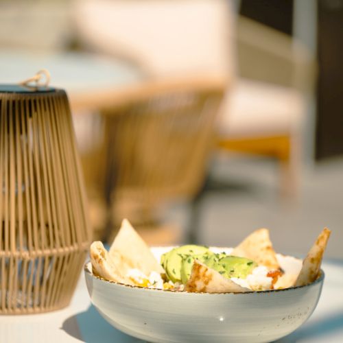 A bowl with tortilla chips, avocado, and cheese sits on a table with a wicker lamp nearby, in an outdoor setting on a sunny day.