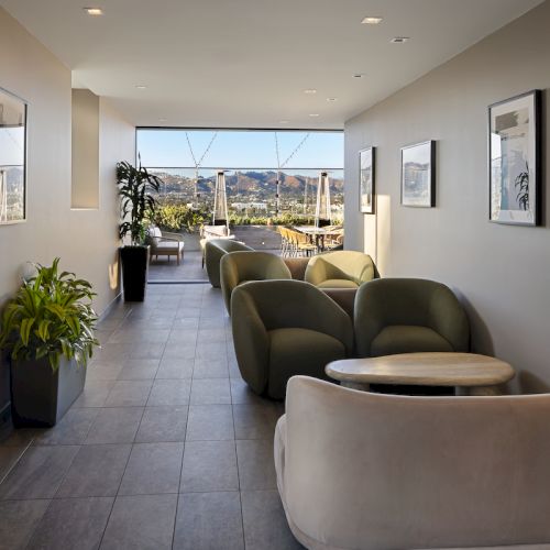 A modern lounge area with cushioned chairs, tables, plants, and artwork on the walls; large windows offer a scenic outdoor view.