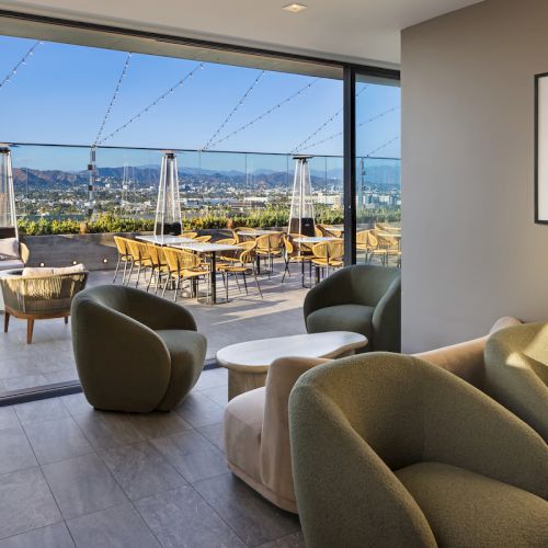 A modern lounge with cozy seating opens to a terrace featuring tables and chairs, overlooking a scenic landscape under a clear sky.
