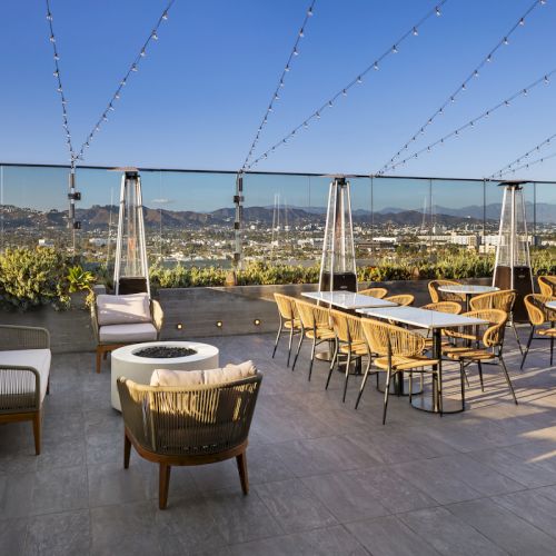 The image shows an outdoor rooftop patio with tables, chairs, and string lights, overlooking a scenic view of a city under a clear blue sky.
