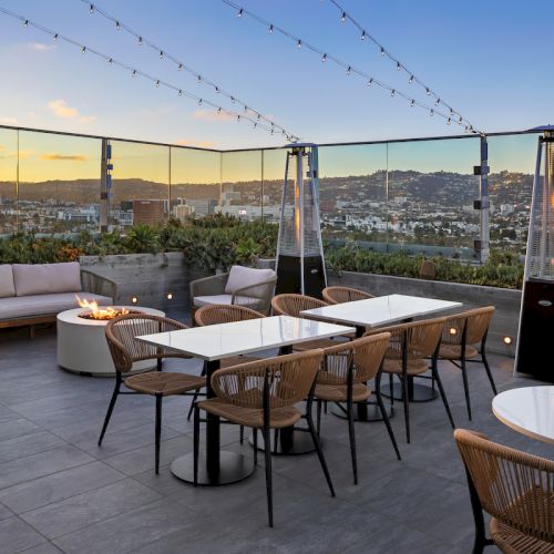 Rooftop patio with tables, chairs, outdoor heaters, and string lights, overlooking a cityscape at sunset.