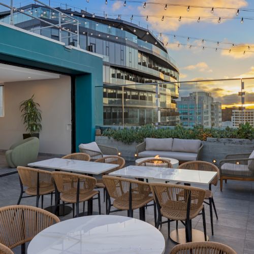 The image shows a stylish rooftop terrace with tables, chairs, string lights, and a sunset view of the city skyline.