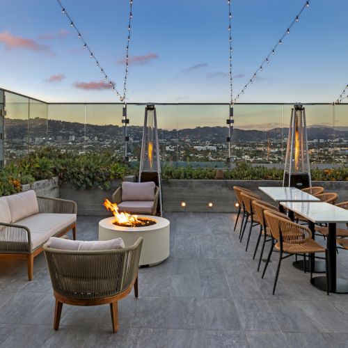 A rooftop patio with seating, tables, a fire pit, and decorative string lights, offering a scenic view of the city at sunset.
