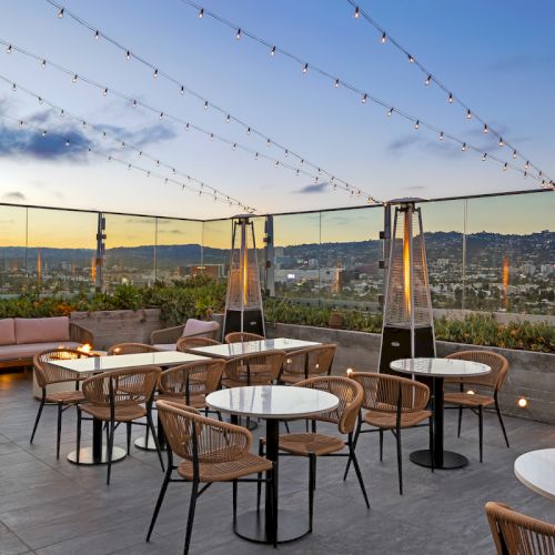 Outdoor patio with tables, chairs, string lights, and heaters; overlooks a scenic view at sunset with a clear sky.